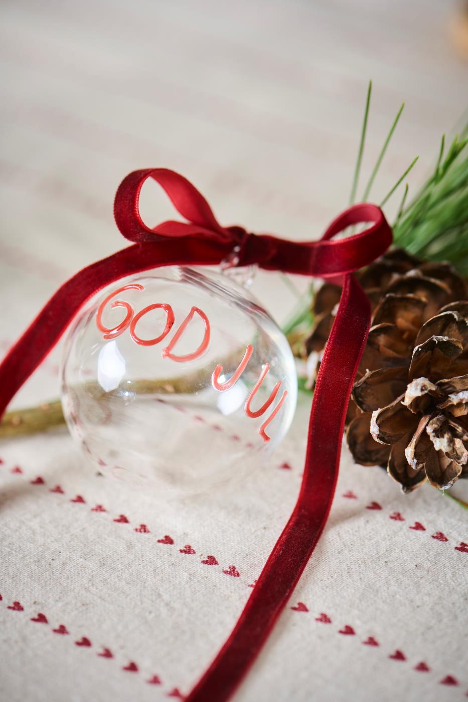 Julgranskula i klart glas med röd text GOD JUL från Ib Laursen liggande på bordet med rosett