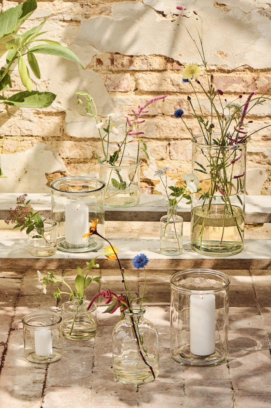 Hannah vaser i klart cylinderformet glas med lys og blomster i på terrasse.