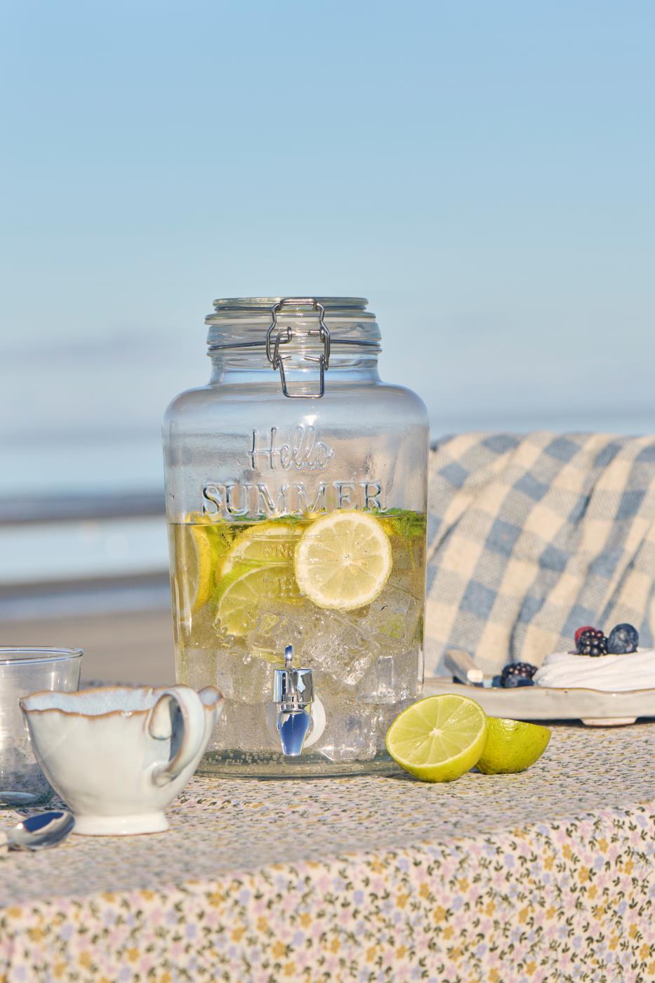 Saftdispenser i klart glas med tappehane fra Ib Laursen stående på bord på stranden